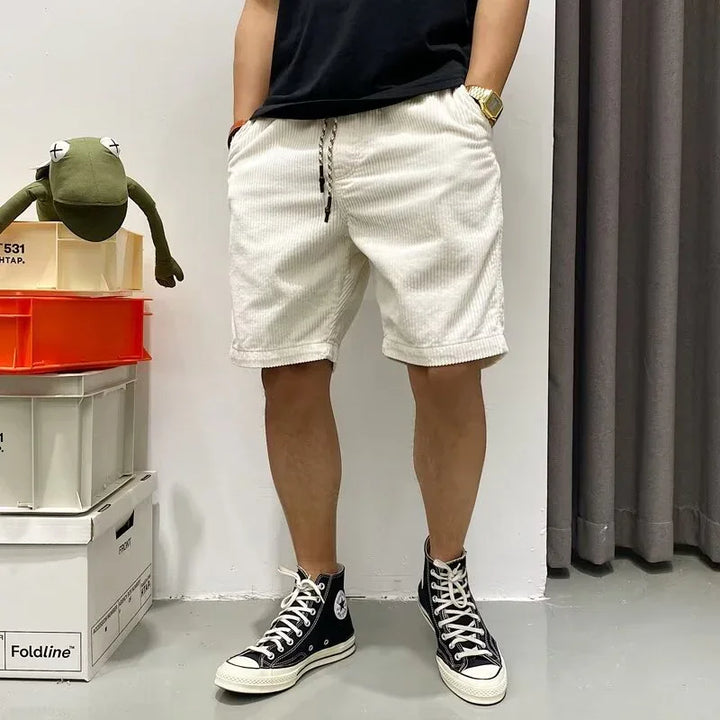 Person wearing white corduroy shorts, black t-shirt, and black high-top sneakers standing indoors