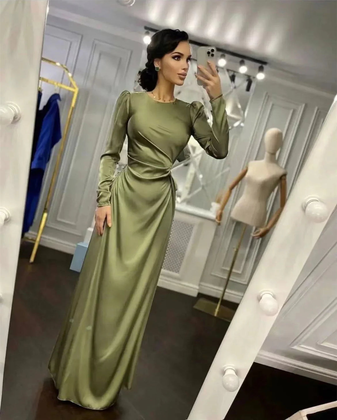Woman in olive green satin evening gown with long sleeves taking mirror selfie in boutique