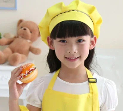 Smiling girl in yellow chef hat and apron holding decorated burger toy indoors with teddy bear background