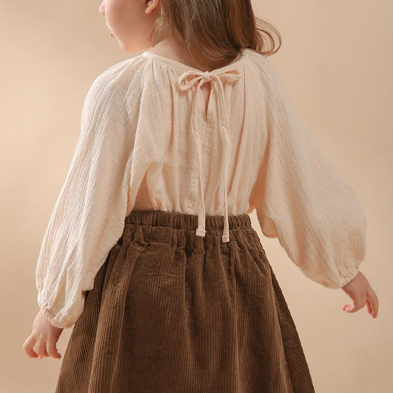 Child wearing cream-colored long-sleeve blouse with back tie and brown corduroy skirt