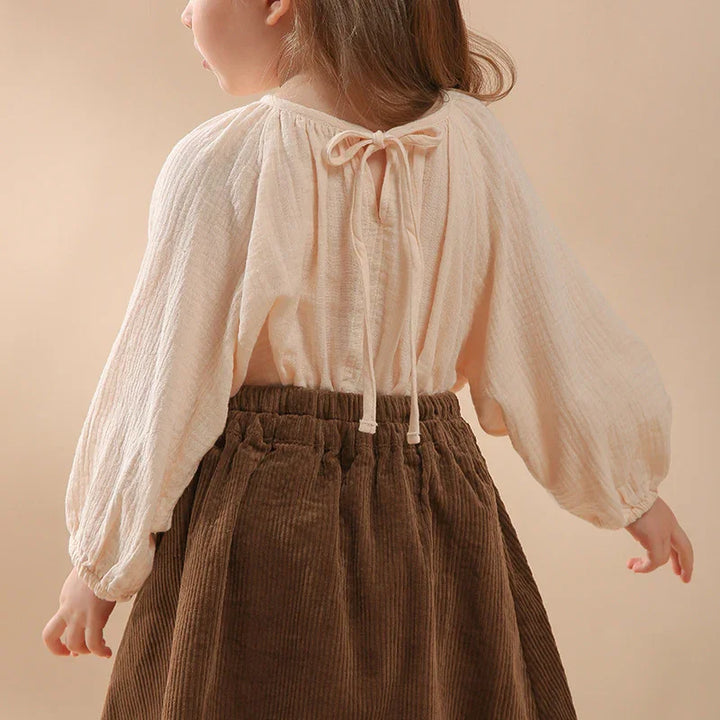 Child wearing cream-colored long-sleeve blouse with back tie and brown corduroy skirt