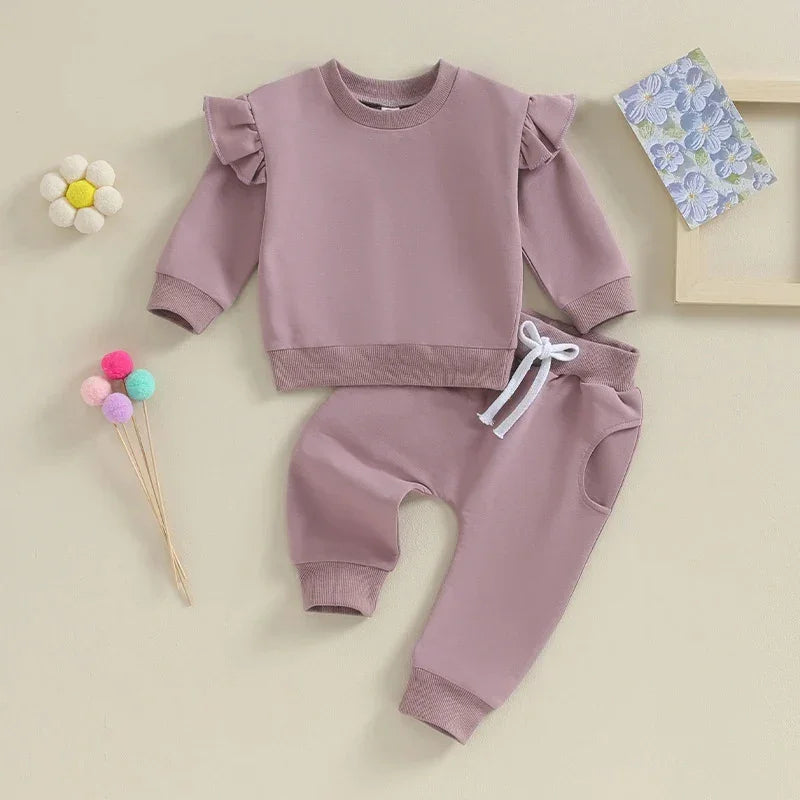 Mauve toddler outfit with ruffled shoulder sweatshirt and drawstring pants on beige background