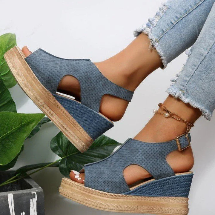Blue wedge sandals with cutout design on feet, paired with light ripped jeans and anklet, green plant background