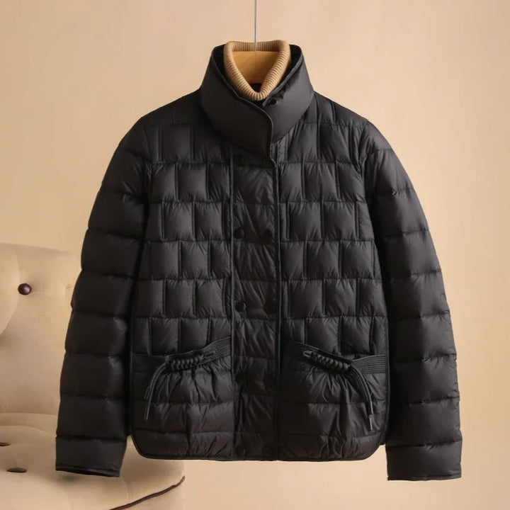 Black quilted puffer jacket with high collar and drawstring pockets on beige chair background