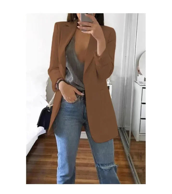 Woman wearing brown blazer with gray top and ripped blue jeans taking mirror selfie indoors