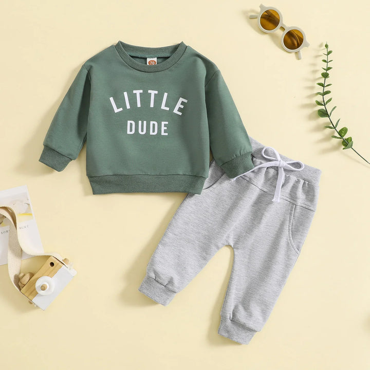 Toddler outfit with green sweatshirt labeled Little Dude and gray sweatpants on beige background