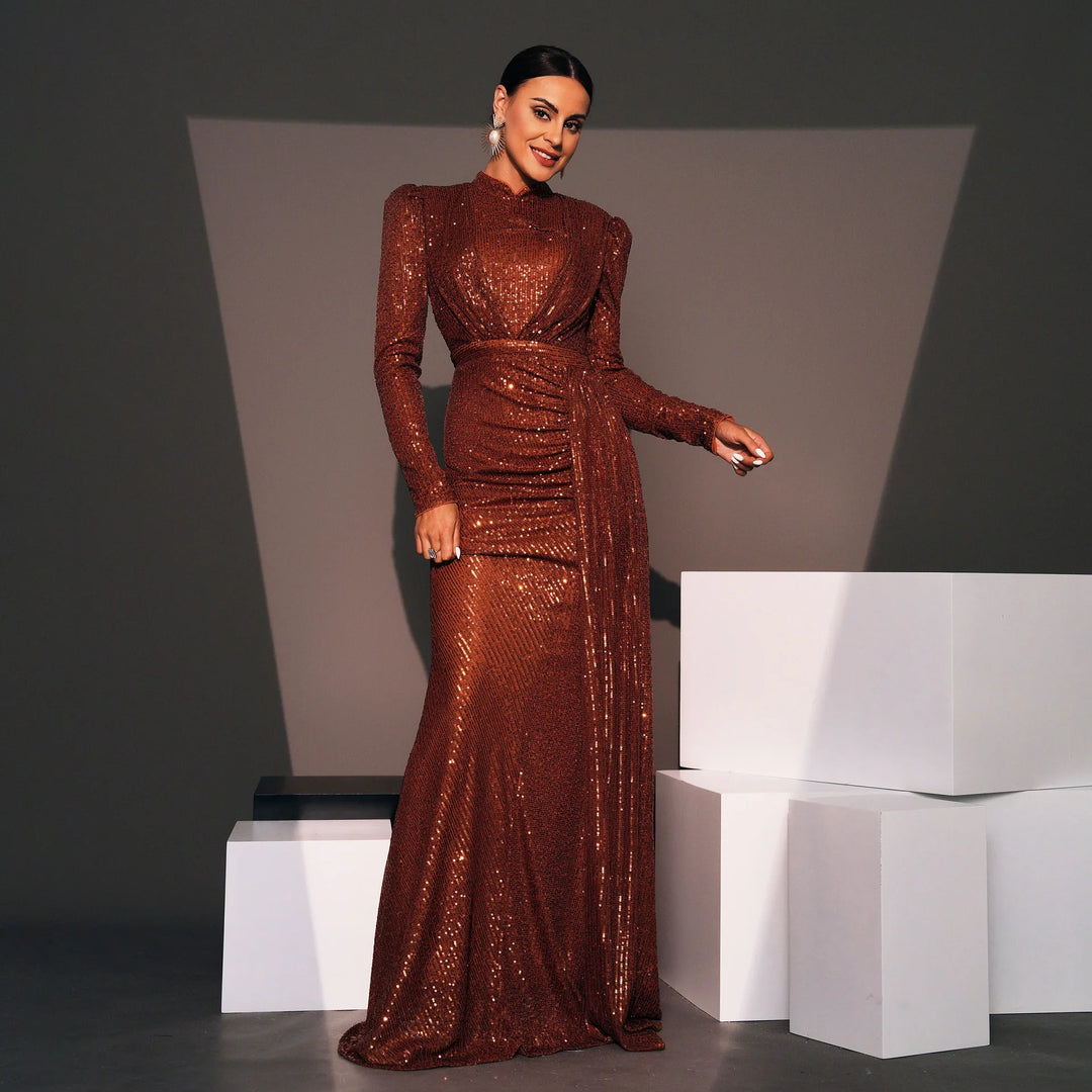 Woman in long, copper sequin gown with long sleeves and draped waist posing against gray backdrop