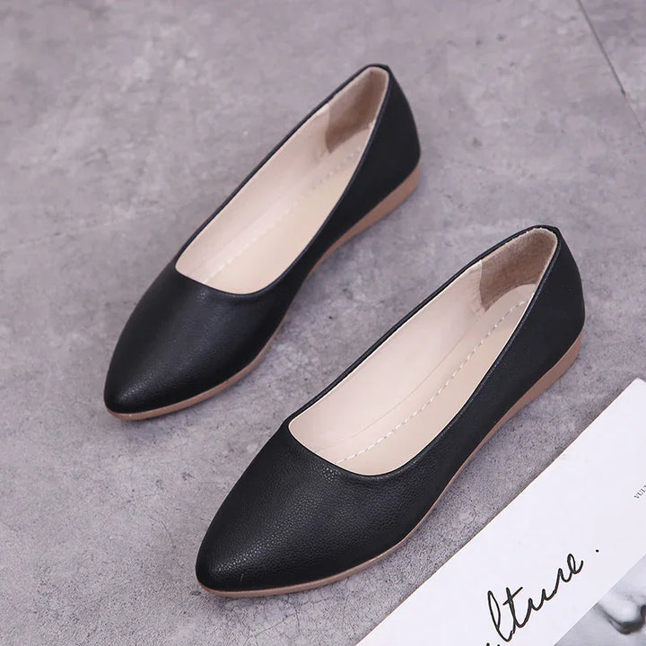 Pair of black pointed-toe flat shoes on a grey surface next to a white paper with cursive text