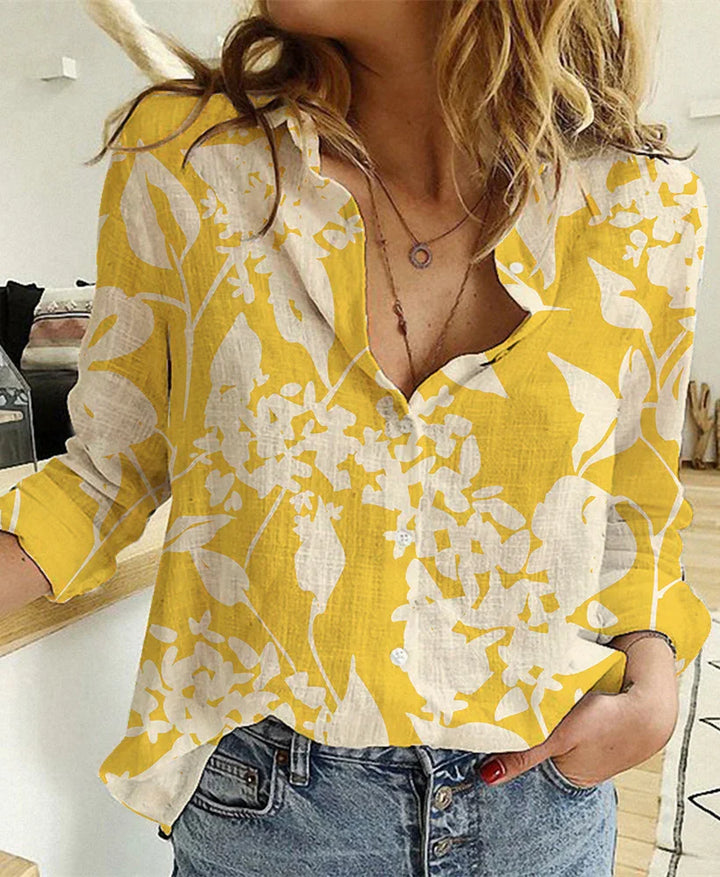 Woman wearing a yellow floral button-up blouse with white leaf patterns and blue jeans indoors