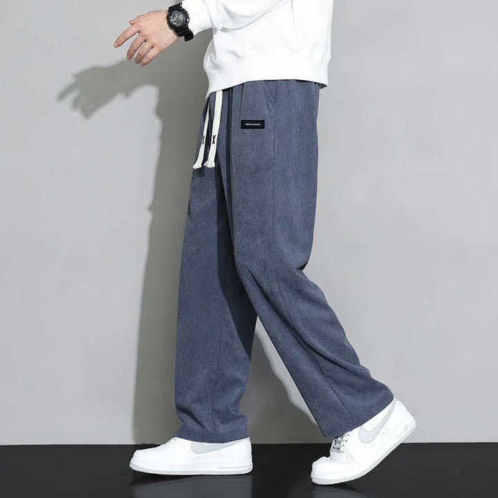 Man wearing blue wide-leg trousers with white drawstrings and white Nike sneakers against gray background