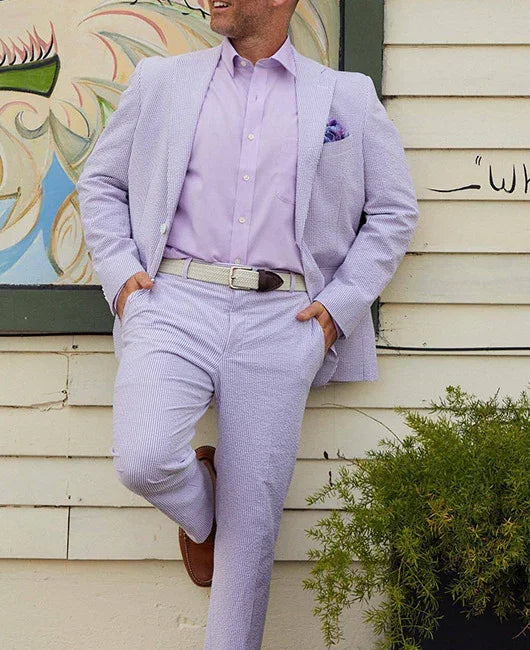 Man wearing light purple seersucker suit with matching shirt and beige belt leaning against wall outdoors