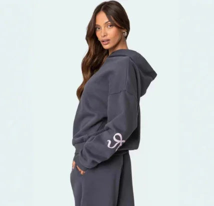 Woman wearing dark gray cozy hoodie and matching sweatpants with logo on sleeve