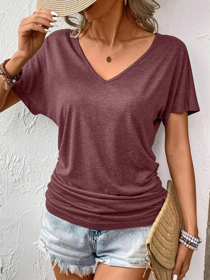 Woman wearing a casual purple V-neck t-shirt with light denim shorts, holding a woven clutch and straw hat