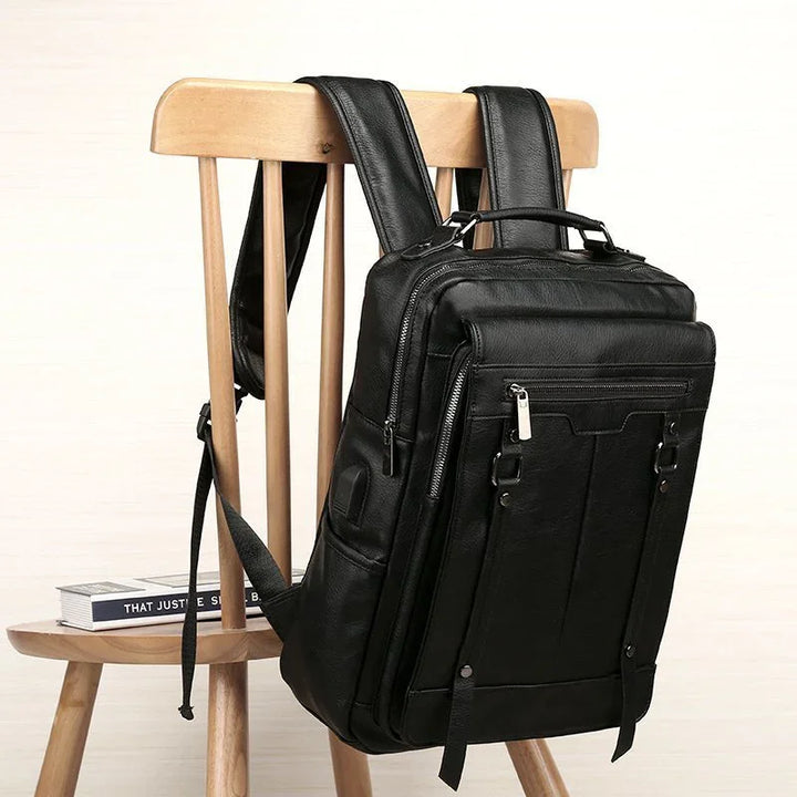 Black leather backpack with multiple zippered pockets hanging on a wooden chair with stacked books