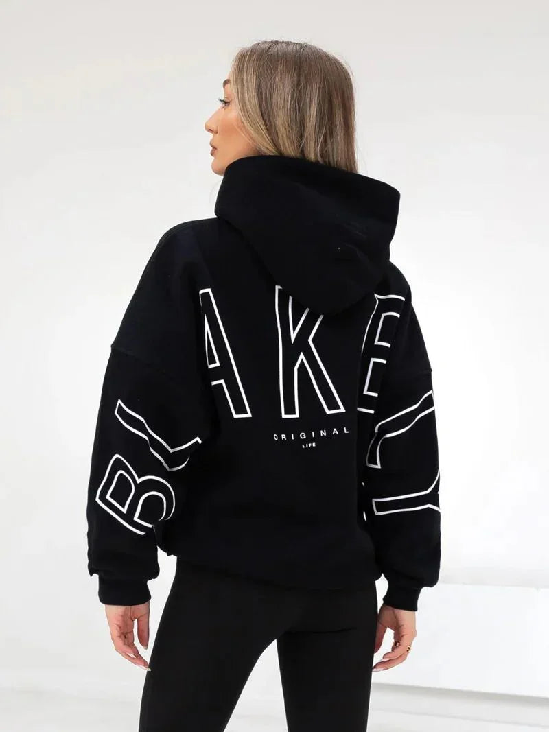 Woman wearing black oversized hoodie with bold white 'BAKED' text and black leggings in minimalist white studio