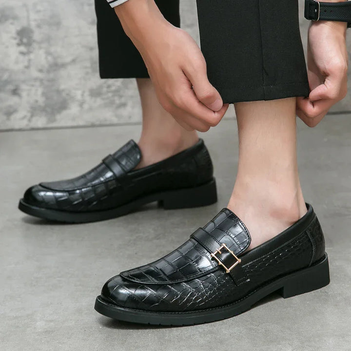 Black crocodile pattern leather loafers with gold buckle detail worn with black trousers