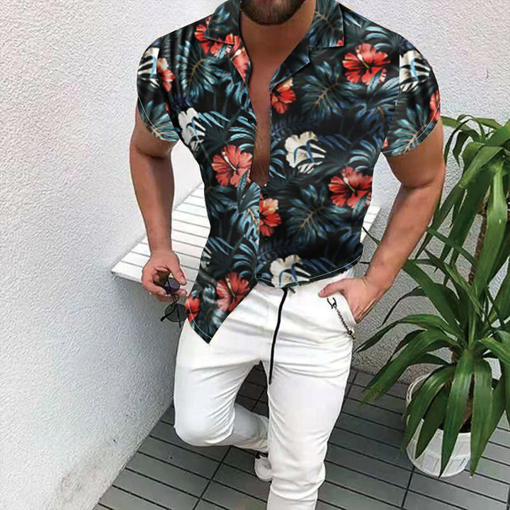 Man wearing black floral short-sleeve button-up shirt with white pants standing near potted plant on tiled floor