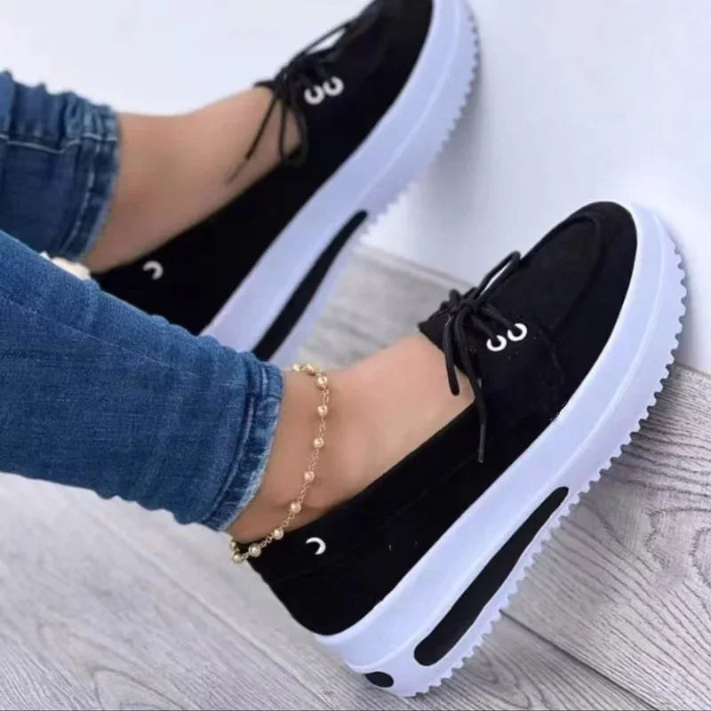 Black casual platform sneakers with white soles worn with blue jeans and gold anklet on wood floor