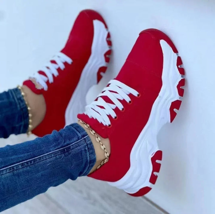 Red and white chunky sneakers with white laces worn with blue jeans and gold anklet on white background