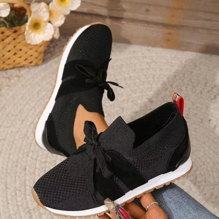 Black knit sneakers with white soles and red heel tabs worn indoors on carpet with basket and flowers