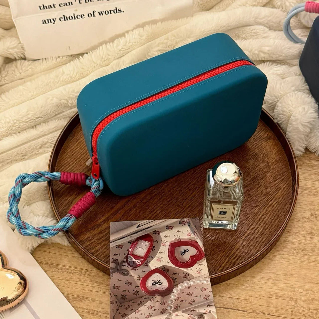 Teal rectangular pouch with red zipper and braided handle, perfume bottle, and photo on wooden tray