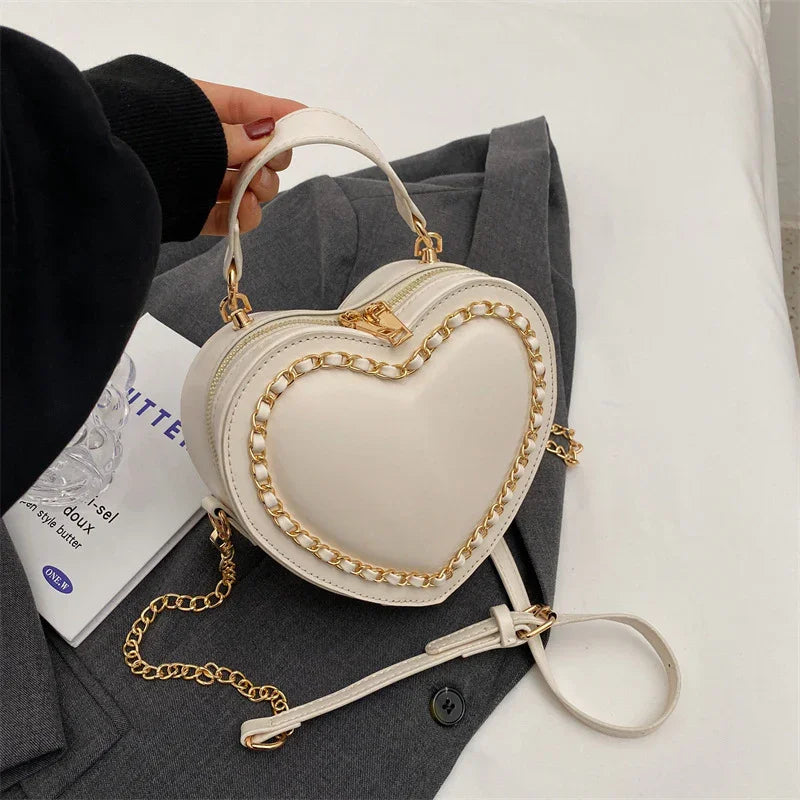 White heart-shaped handbag with gold chain detail and top handle on gray coat