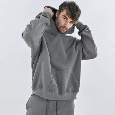 Man wearing a gray oversized hoodie and matching pants pulling up the hood against a plain white background