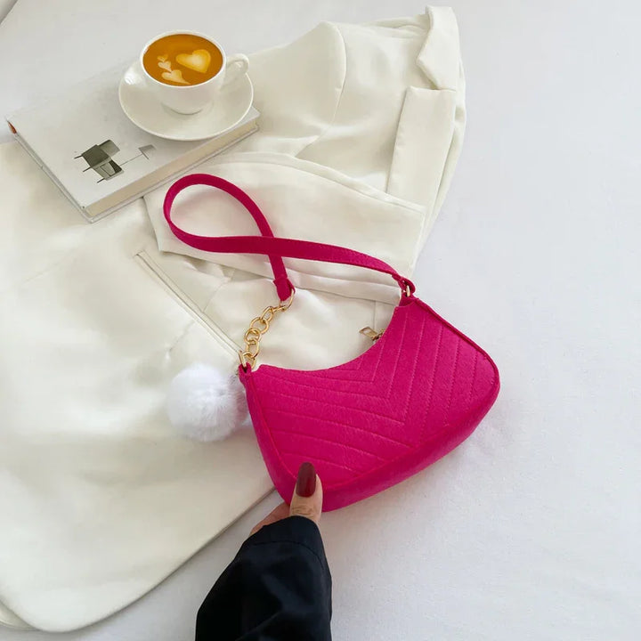 Hand holding vibrant pink quilted handbag with gold chain and white pompom on white fabric and cup of coffee