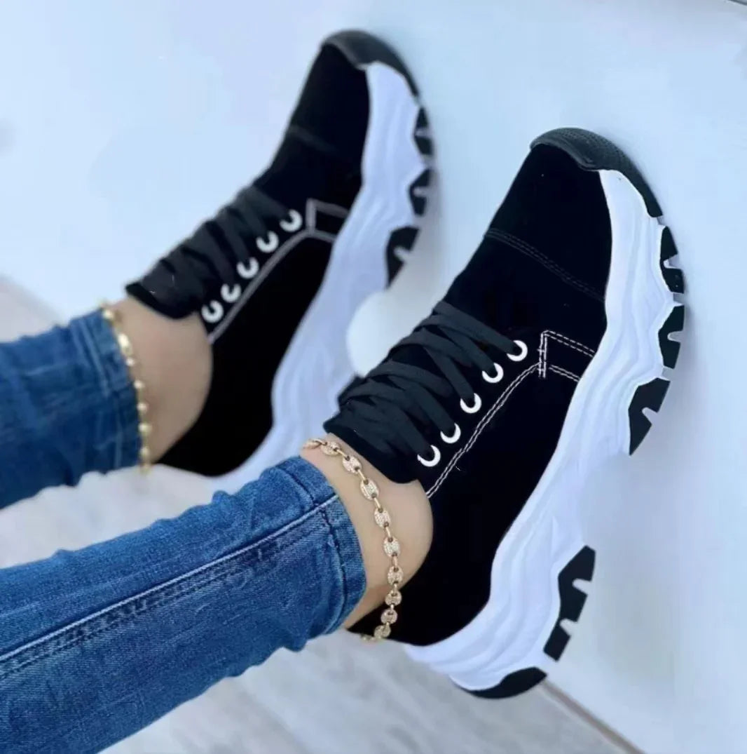 Black chunky sole sneakers with white stitching and thick white patterned outsole worn with blue jeans and gold anklets