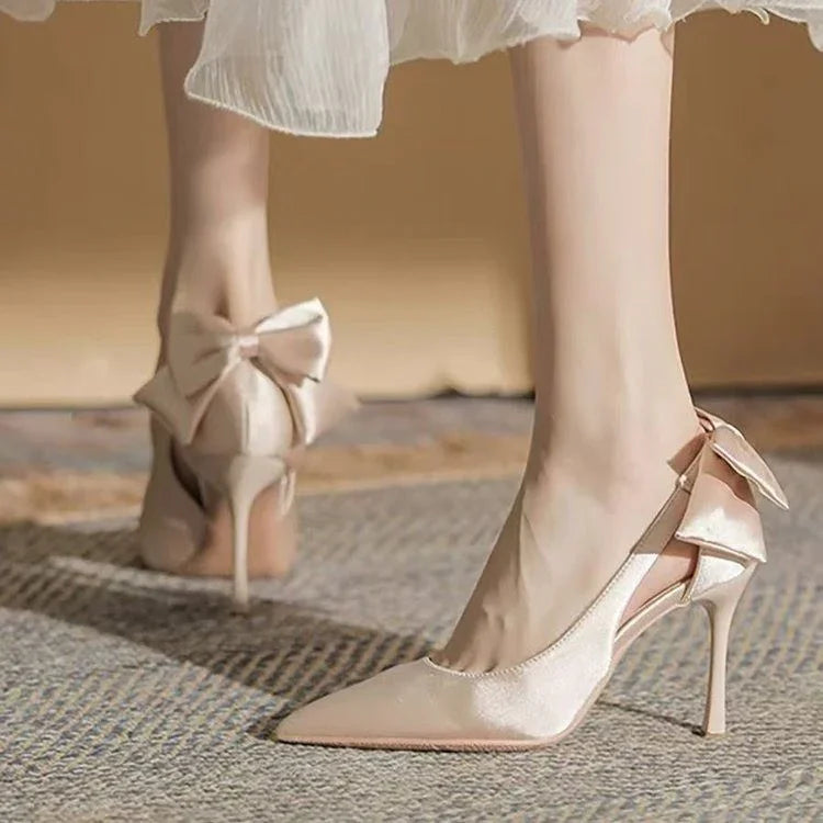 Elegant beige satin high heels with pointed toe and decorative bows on back, worn with white dress