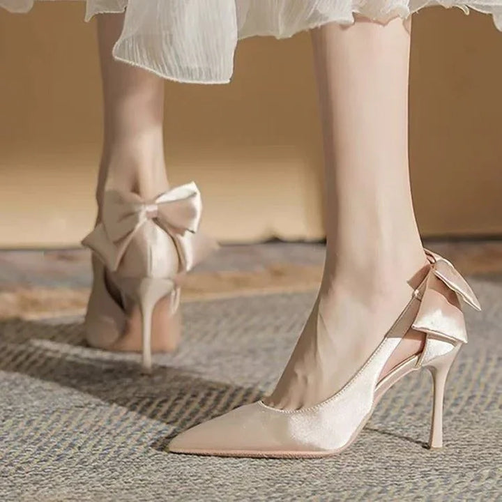 Elegant beige satin high heels with pointed toe and decorative bows on back, worn with white dress