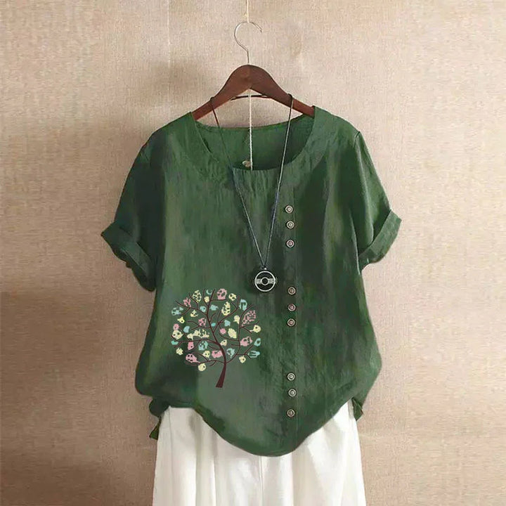 Green short-sleeve blouse with buttons and floral tree print, paired with white pants and pendant necklace