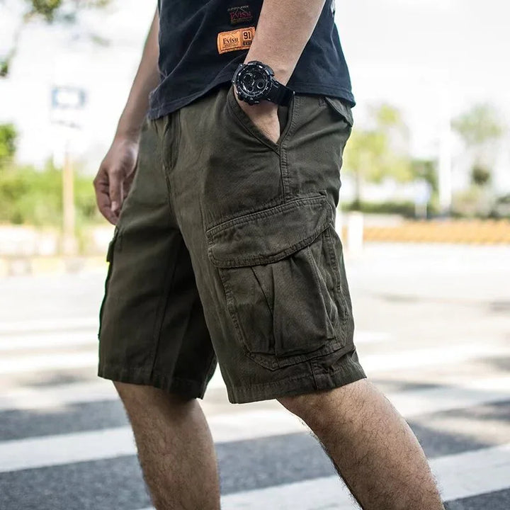 Man wearing olive green cargo shorts and a black watch standing outdoors urban setting