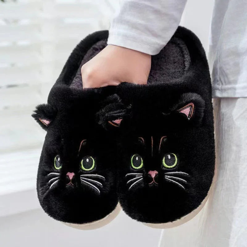 Hand holding black plush slippers with cute green-eyed cat face and ears, cozy indoor footwear