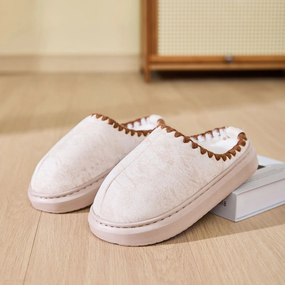 Pair of beige cozy indoor slippers with brown trim on wooden floor near a book