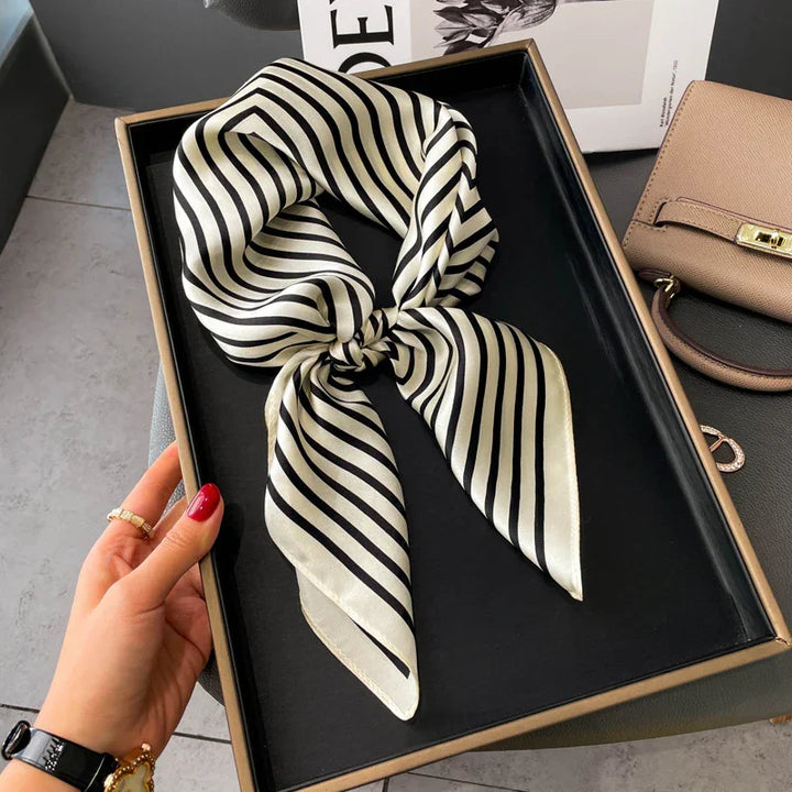 Black and white striped silk scarf displayed in black gift box on table with beige handbag nearby