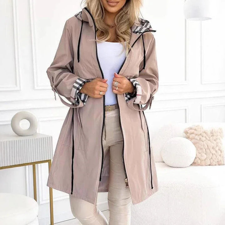Woman wearing beige long jacket with plaid cuffs and hood, white top, and light pants in modern room