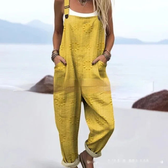 woman wearing yellow textured overalls with rolled cuffs standing on a beach with rocks and mountains in background