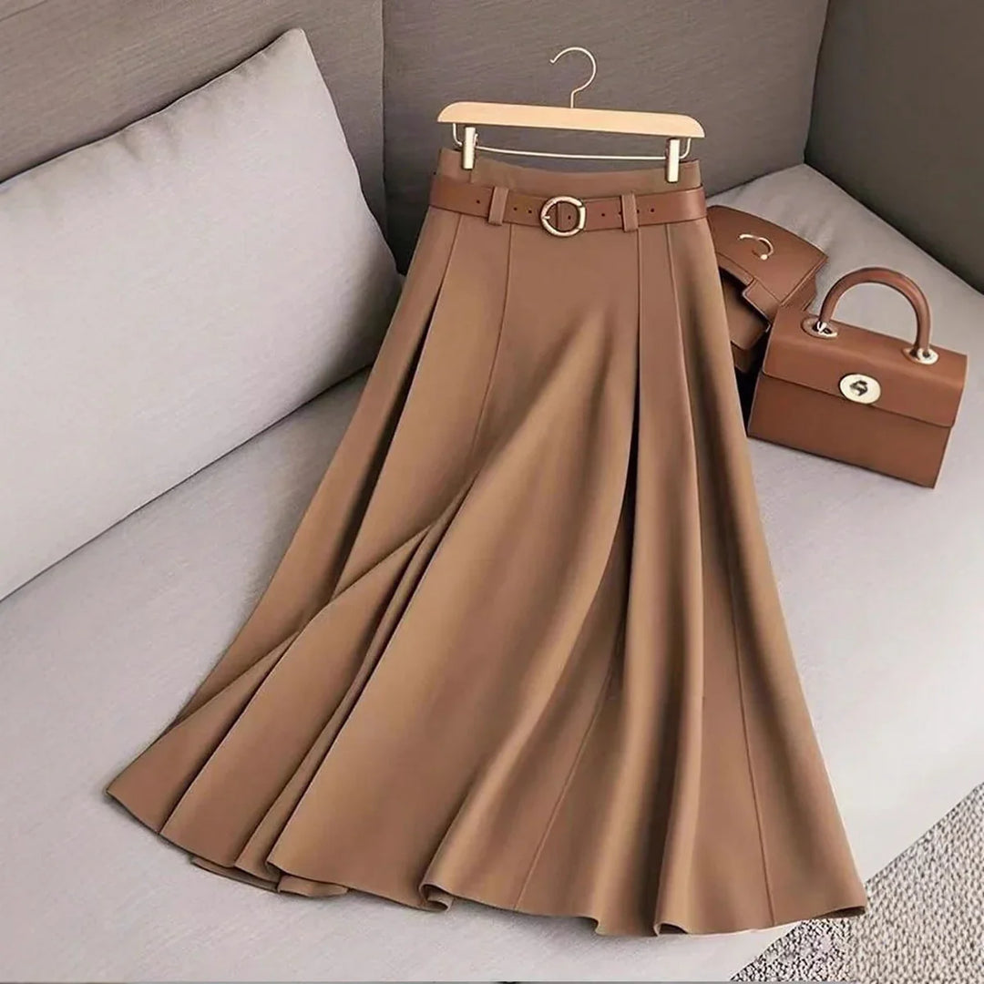 Brown pleated midi skirt with matching belt and gold buckle, displayed on gray sofa with tan handbag