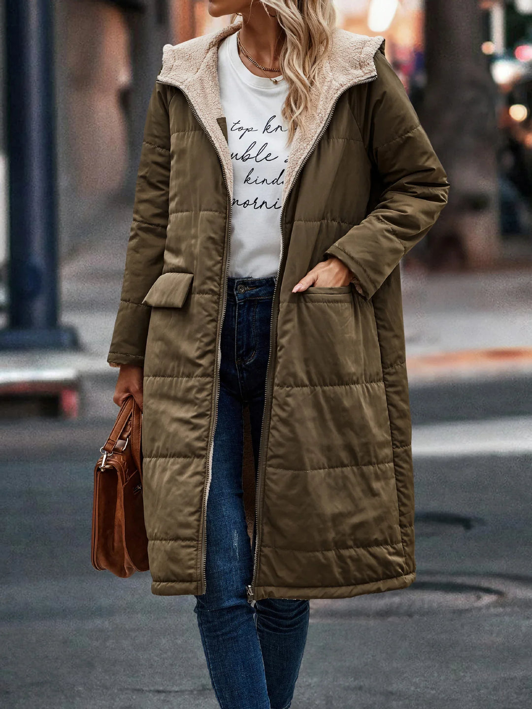 Woman wearing long olive green quilted coat with fleece lining over casual white graphic tee and jeans, city street background