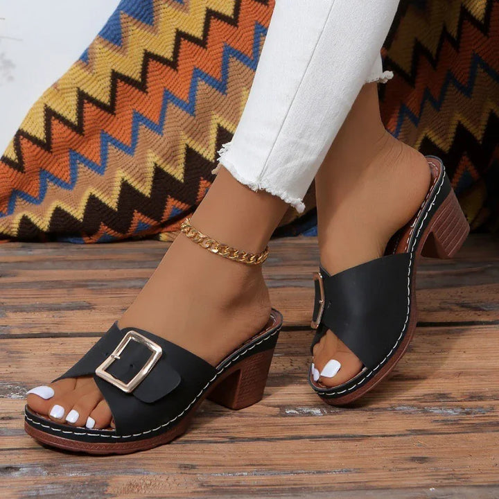 Black leather heeled mules with buckle detail worn with white jeans and gold anklet on wooden floor