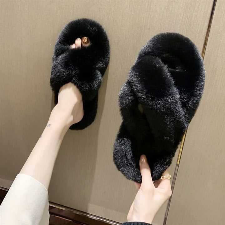 Pair of black fuzzy slippers with crisscross straps worn and held by a person against a beige background
