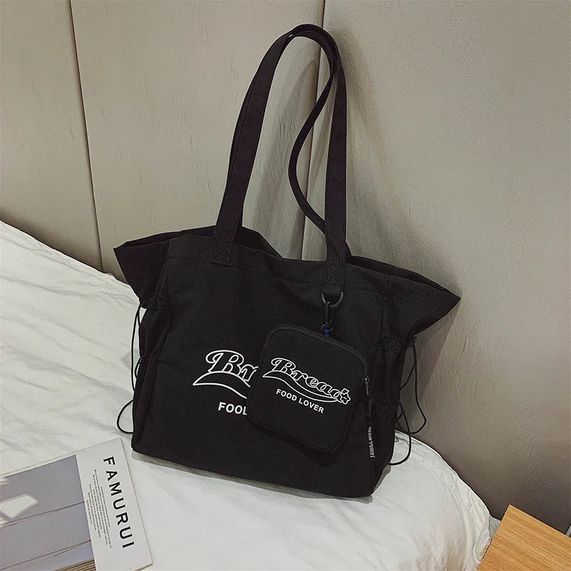 Black fabric tote bag with matching pouch, white graphic text, resting on bed next to a magazine