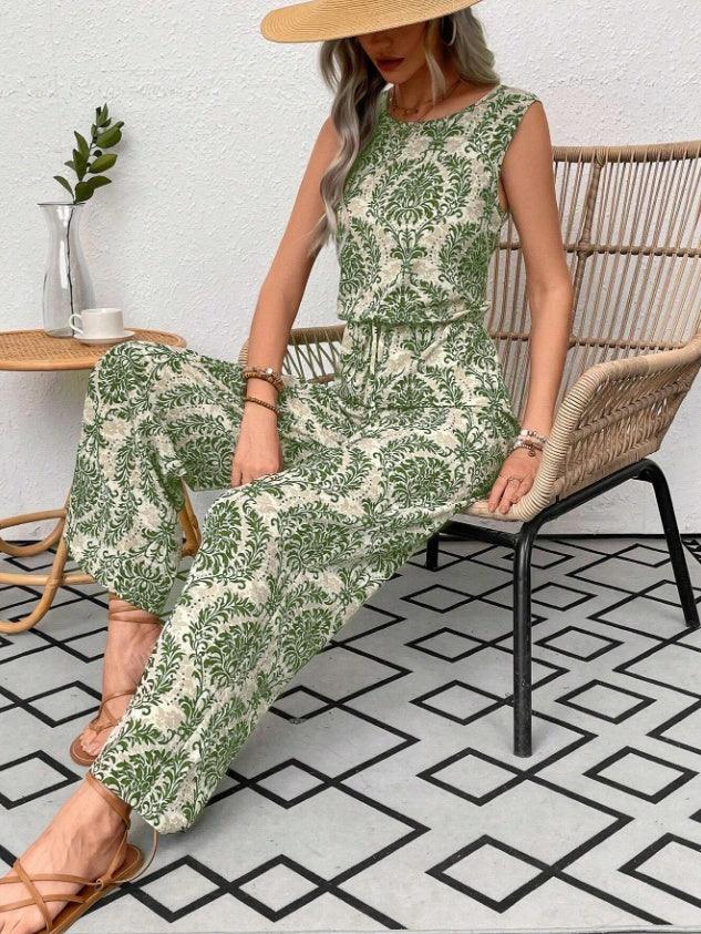 Woman wearing a sleeveless green and beige floral jumpsuit with wide legs, sitting on a wicker chair indoors