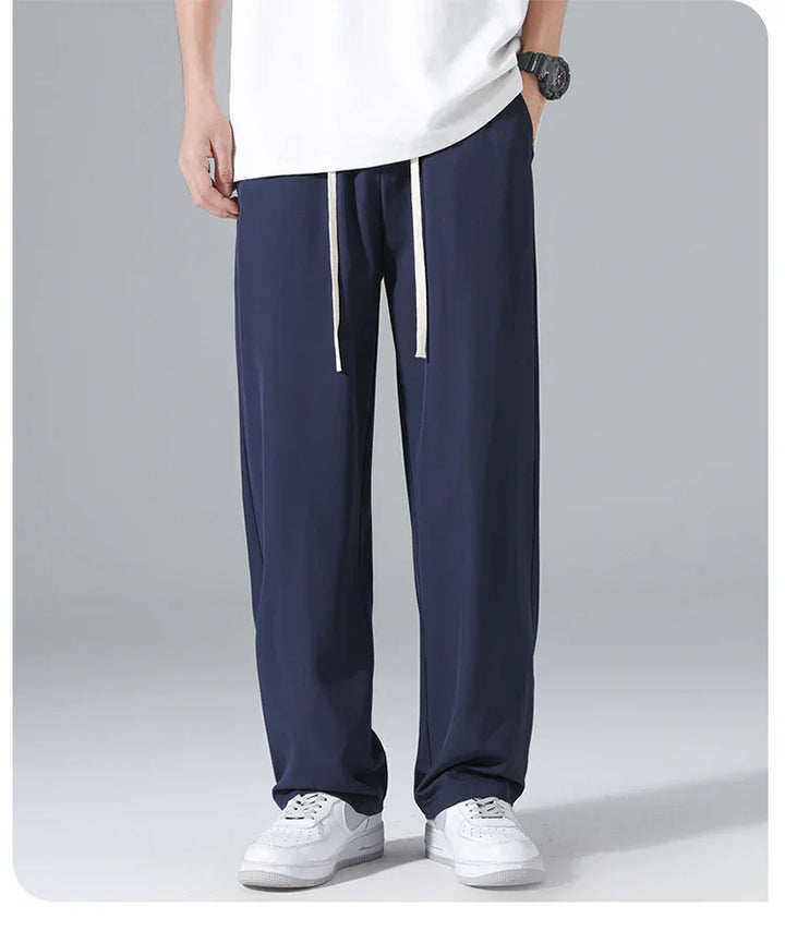 Man wearing navy blue casual pants with white drawstrings and white sneakers, standing against gray background