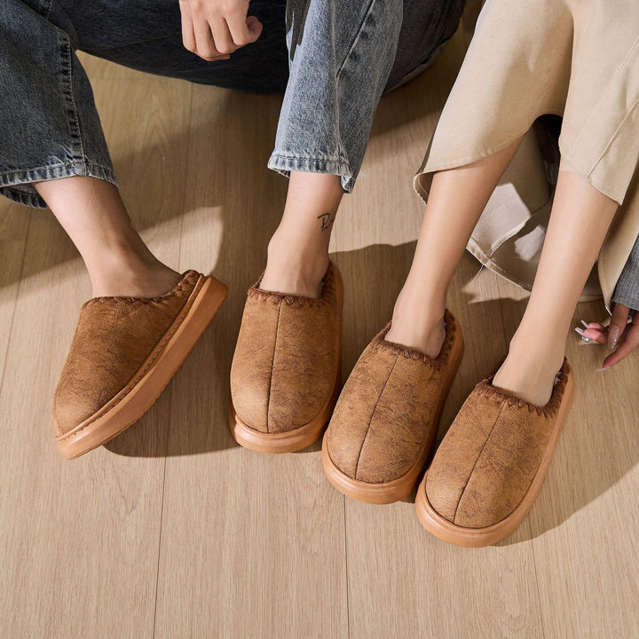 Two pairs of brown cozy slippers worn by people sitting on wooden floor indoors