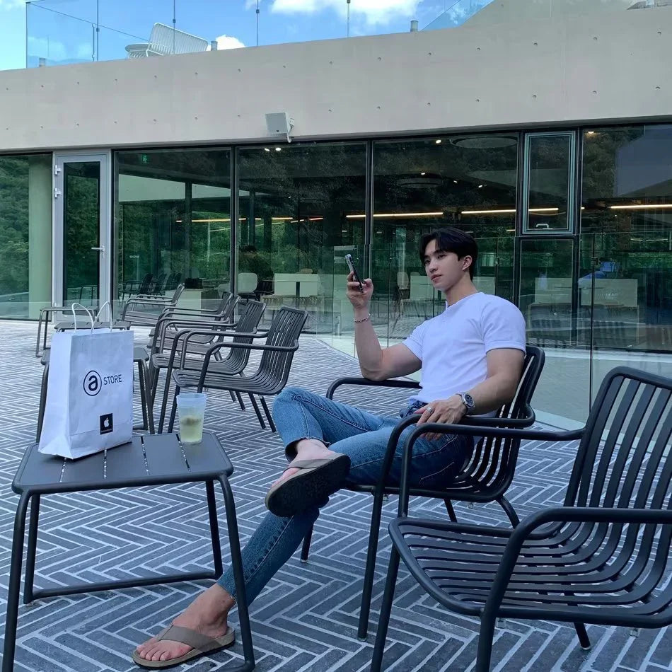 Young man in white t-shirt and jeans sitting on outdoor black metal chair using smartphone at modern cafe patio