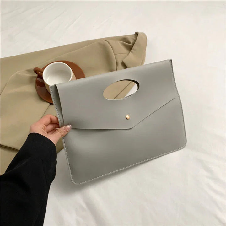 Hand holding sleek gray leather envelope clutch bag with beige coat and ceramic cup on brown saucer in background