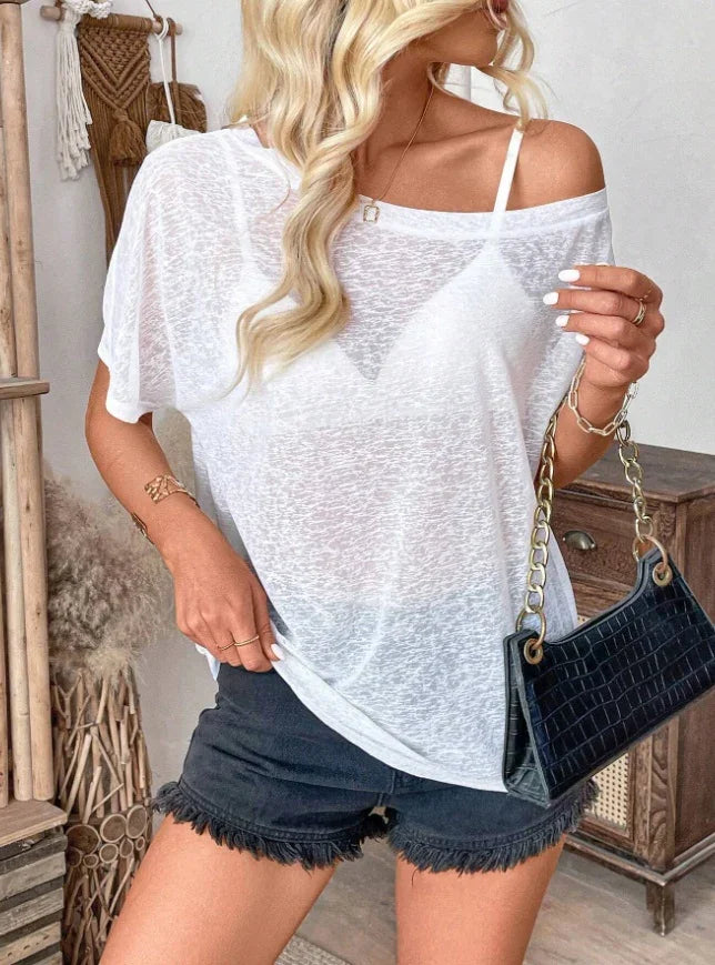 Woman wearing a sheer white off-shoulder top with black bra, black frayed shorts, holding a black handbag with gold chain in boho room