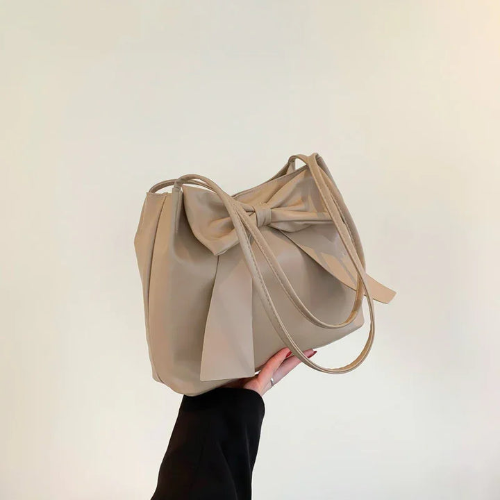 Hand holding beige leather tote bag with large bow detail against plain background